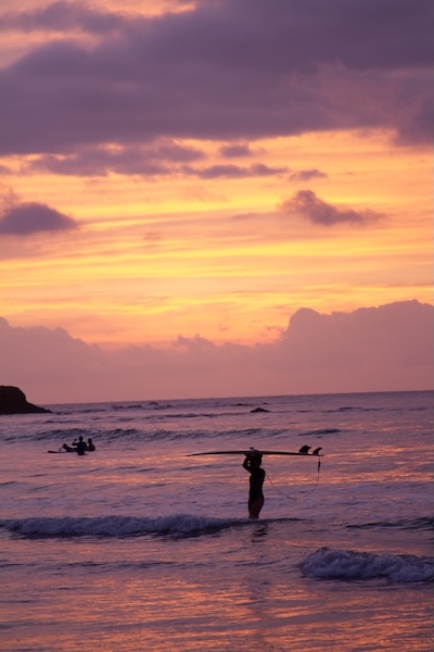 Femme tenant une planche de surf au-dessus de la tête dans l'eau au coucher du soleil