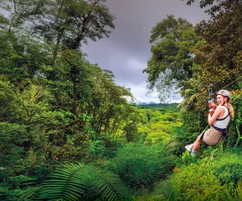 Un voyageur qui descend en tyrolienne à travers la canopée de la dense forêt tropicale costaricaine, suspendu au-dessus d'une végétation luxuriante, avec les montagnes en arrière-plan.