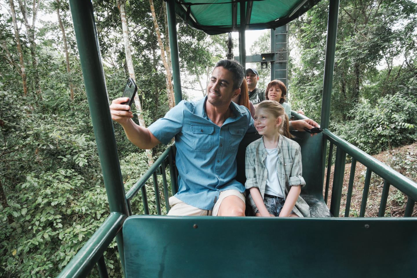 Une famille prend un selfie dans une télécabine en plein air, au-dessus de la canopée de la forêt tropicale.