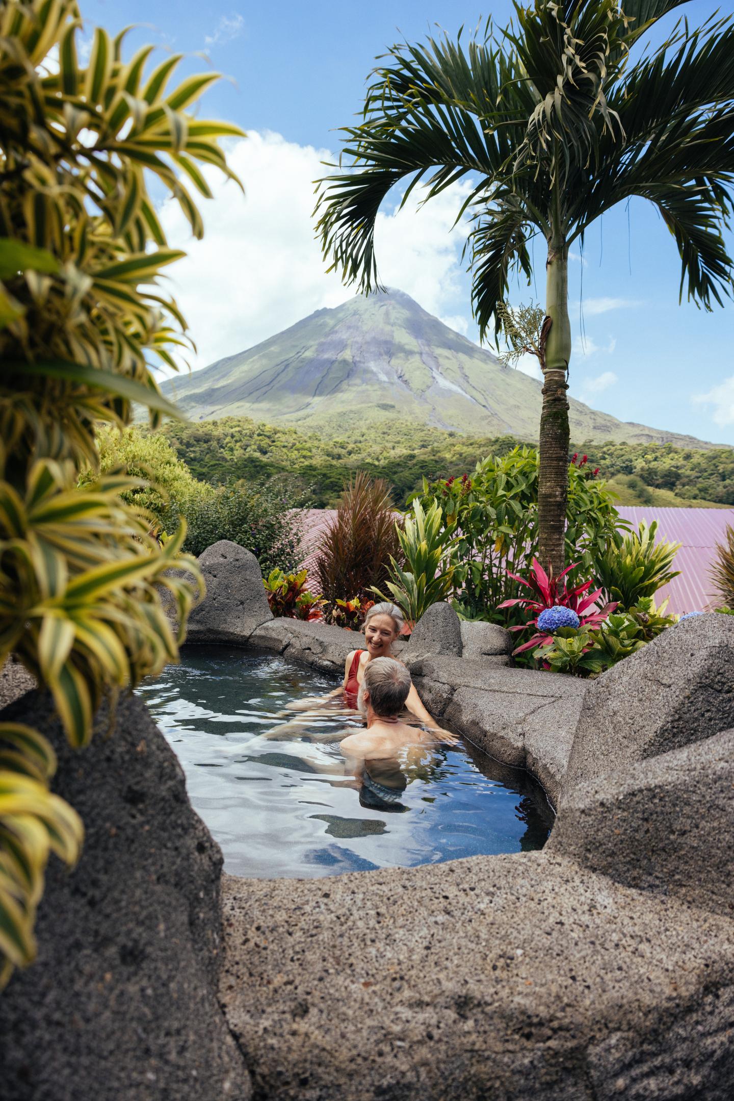 Deux personnes se détendent dans un bassin thermal en pierre, entouré de plantes tropicales, avec le volcan Arenal en arrière-plan.